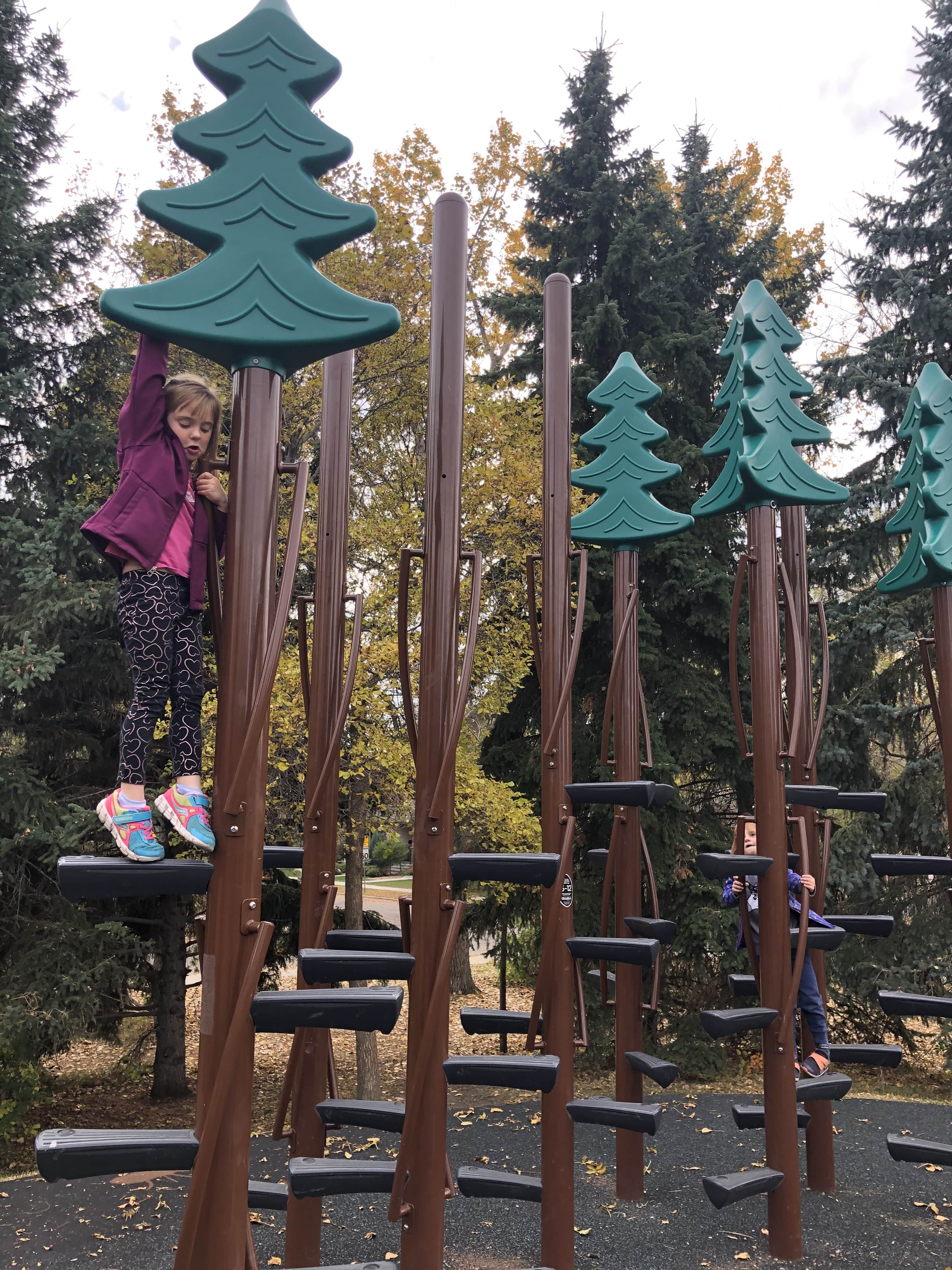Cluster of tall brown climbing poles with staggered black steps and green tree-shaped tops, a person climbing on the left and another on the right.