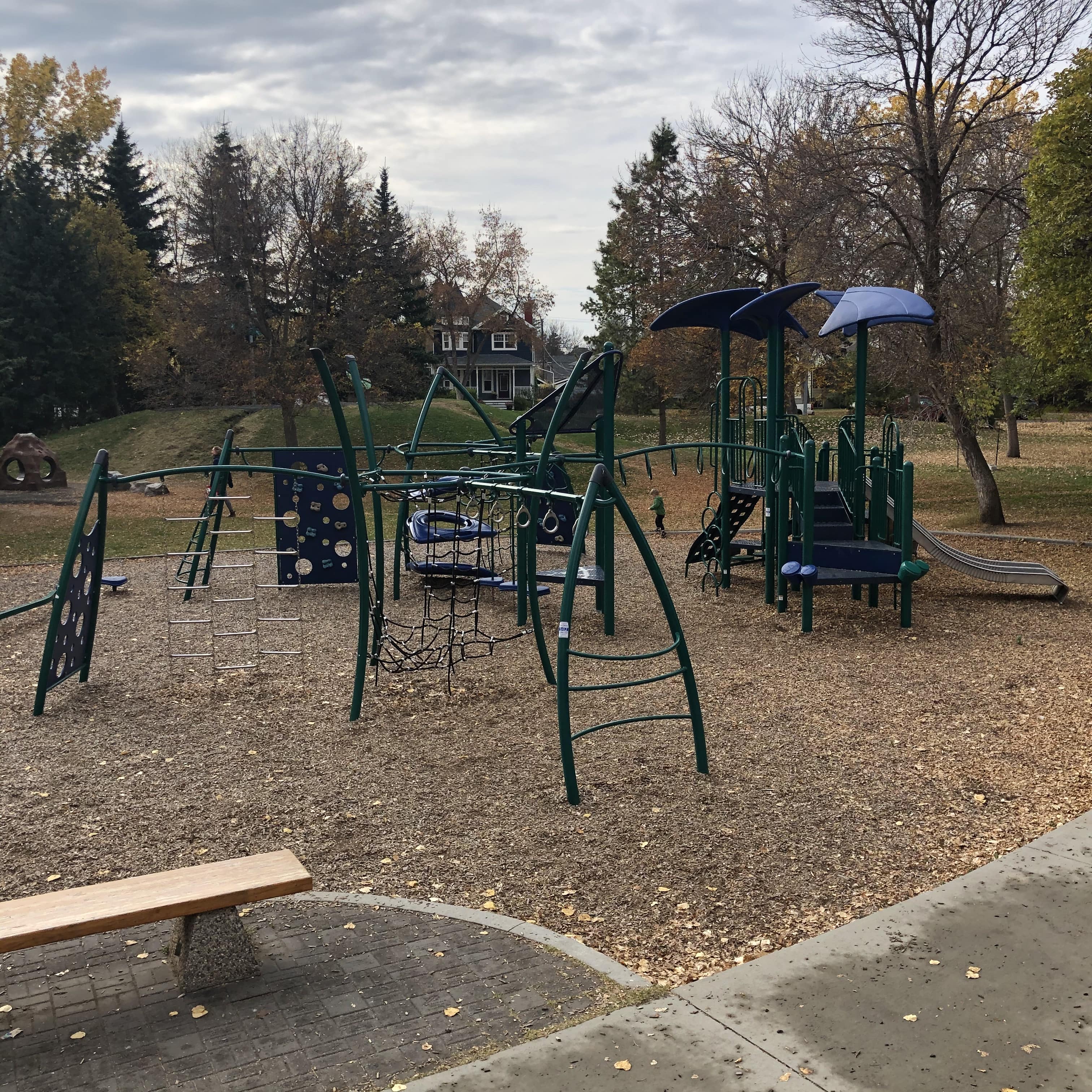 Girl climbing a black rope-and-chain net beside white curved climbing panels and swings on woodchip ground with autumn trees.