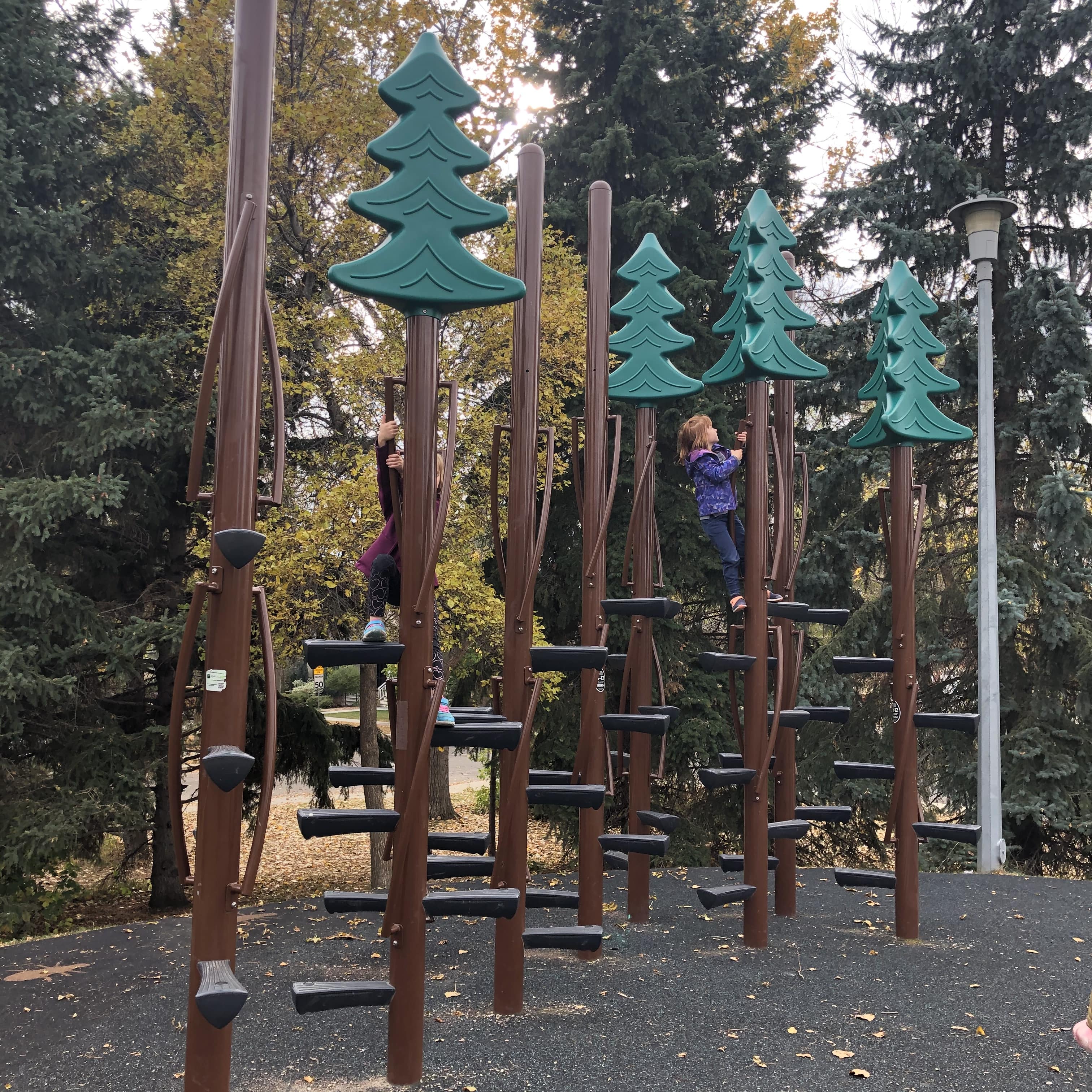 Row of brown vertical poles topped with green stylized tree panels; two people grip poles while climbing beneath evergreen trees and a lamp post.