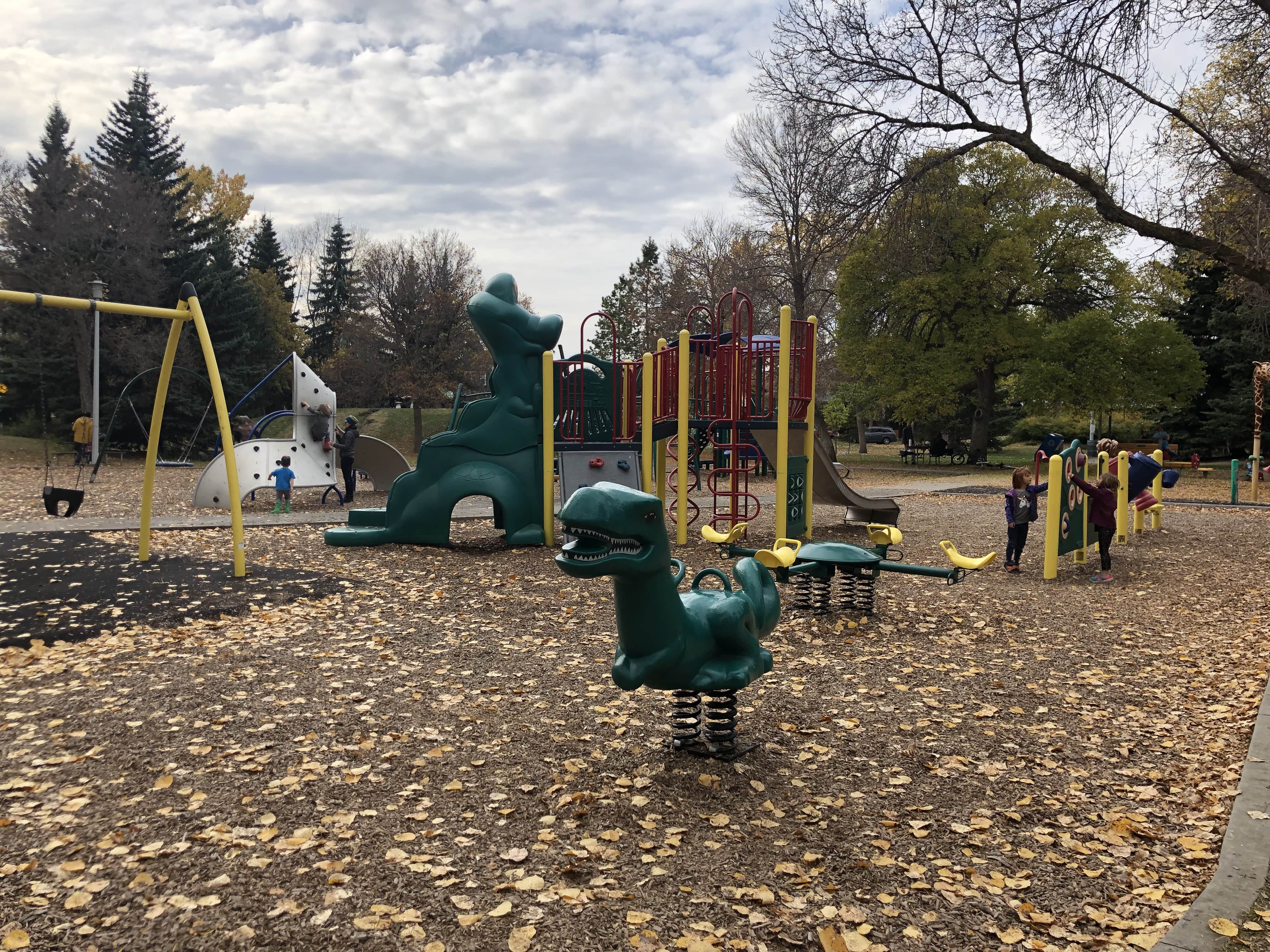 Wide playground scene with a yellow swing frame, a green dinosaur spring rider in the foreground, and a red-and-yellow play structure behind.