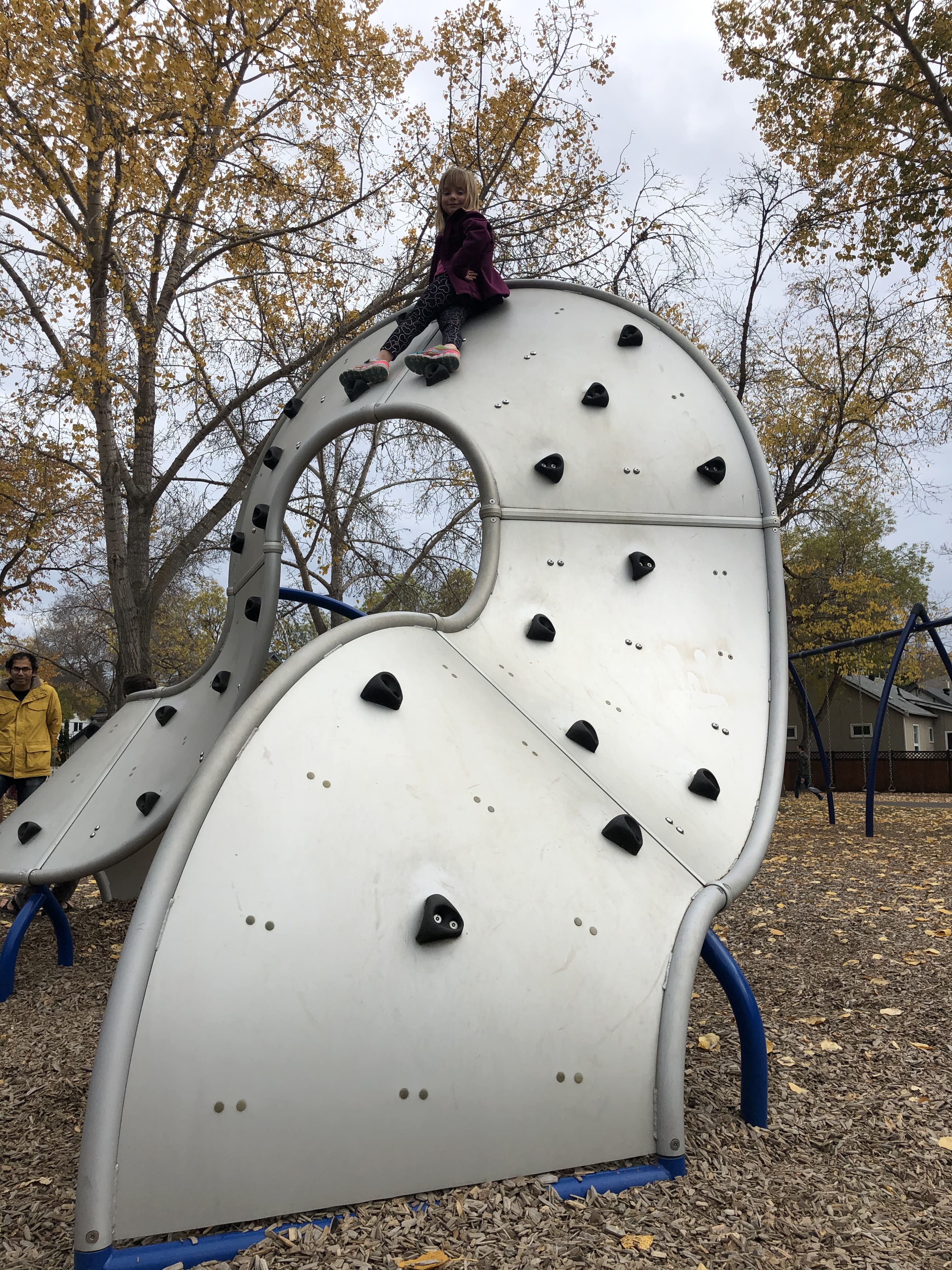 White sculpted climbing wave with scattered black handholds; a child sits at the top against autumn trees in the background.