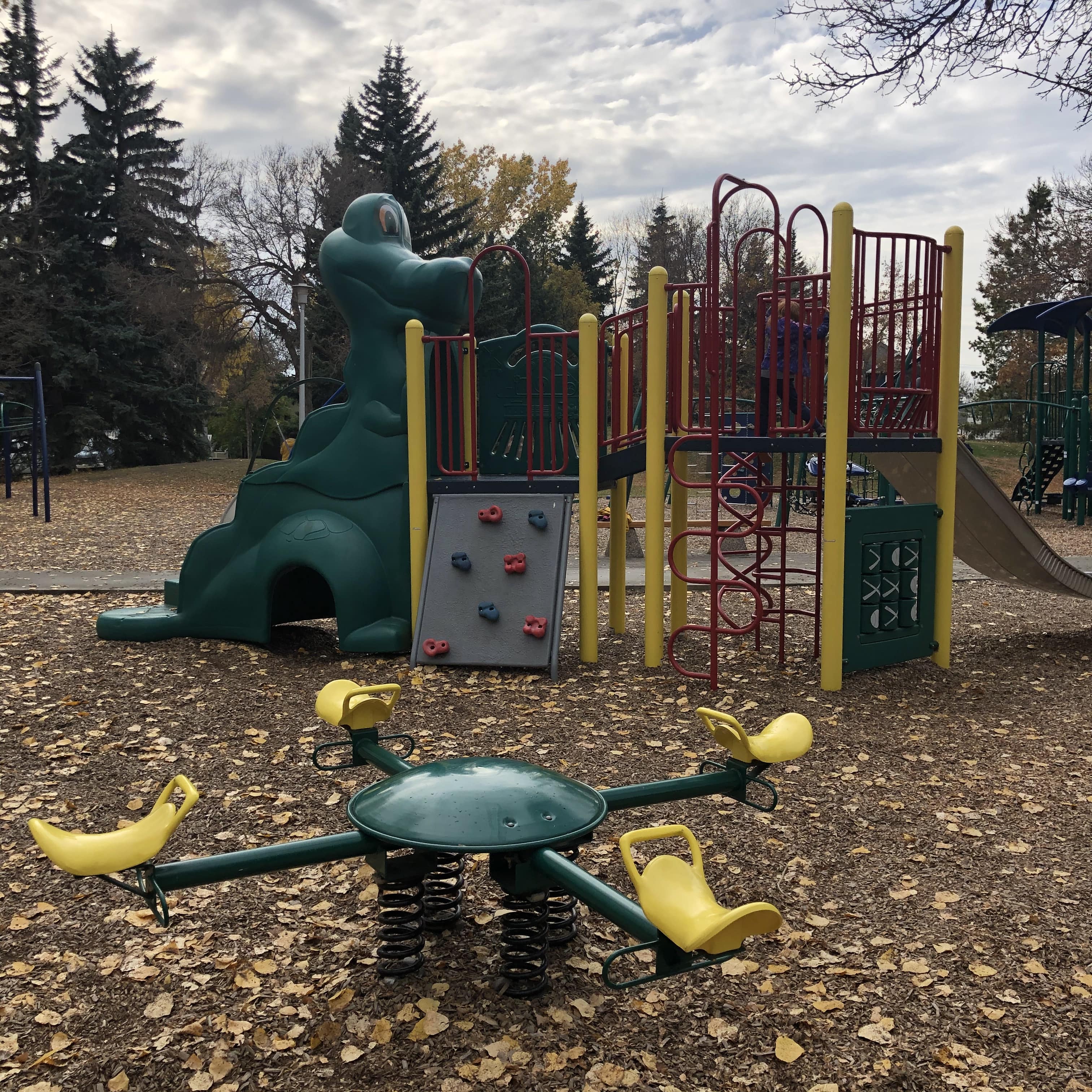 Child climbing a white arched climbing wall with black handholds; picnic table and deciduous trees with fallen leaves behind.