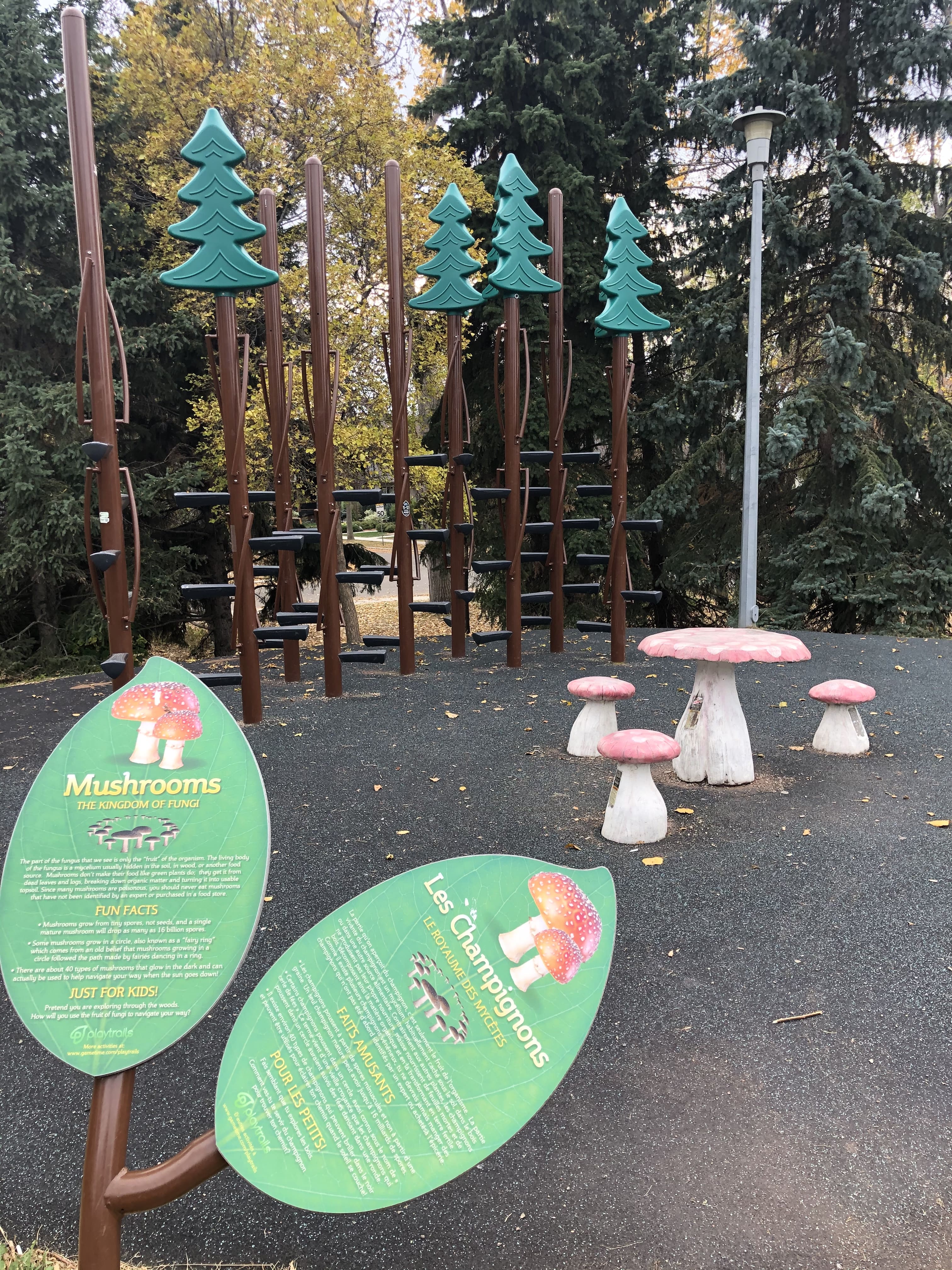 Green interpretive "Mushrooms" sign beside pink toadstool seats and a cluster of brown climbing poles topped with green tree shapes.