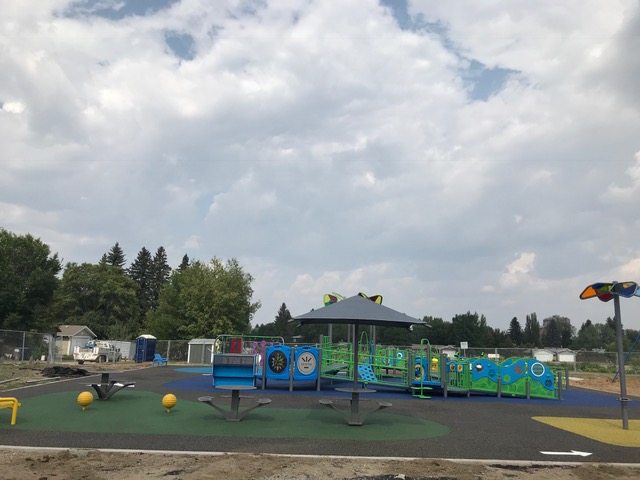 Wide view of playground with central dark shade canopy, green-and-blue play structures and three round spinning platforms on green rubber surfacing.