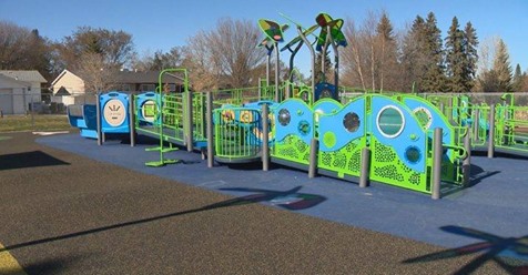 Bright green and blue play structure with wavy panels, circular cutouts, ramps and blue safety surfacing under clear sky.