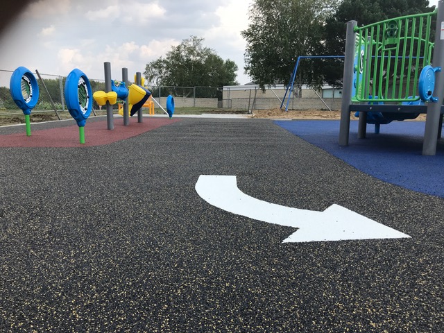 Low-angle view of black rubber path with white directional arrow, nearby blue circular musical panels and yellow play tubes.