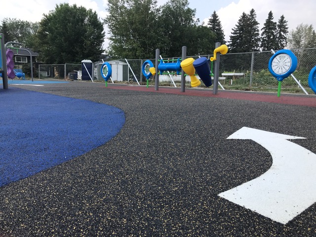 Low-angle view along black and blue rubber path showing white painted arrow and a row of blue circular panels and yellow-and-blue musical tubes by the fence.