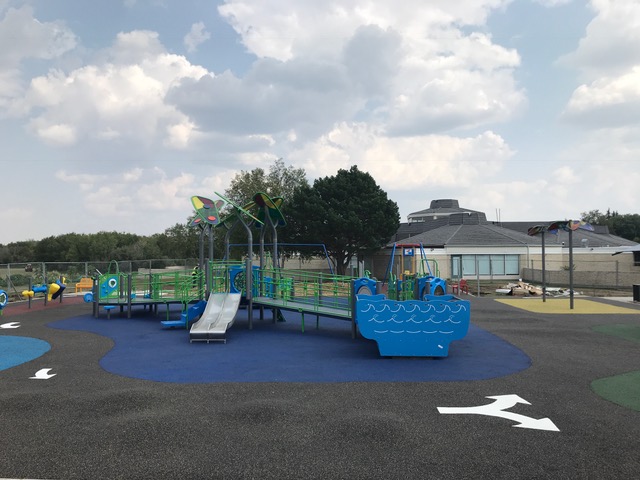 Elevated blue play structure with boat-shaped bow panel, green ramps, twin short slides and leaf-shaped shade canopies under a cloudy sky.