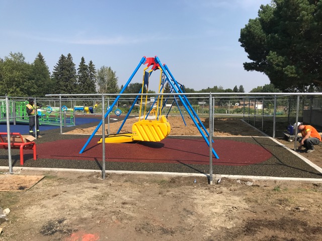 Large yellow molded swing seat suspended in blue frame over red textured surfacing, fenced construction area with workers installing nearby.