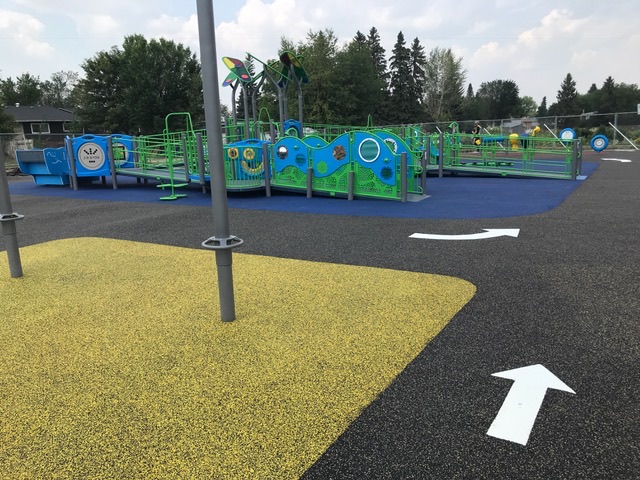 Yellow pad and white directional arrows on dark surfacing; behind, green-and-blue ramped play structure with circular panels and leaf canopies.