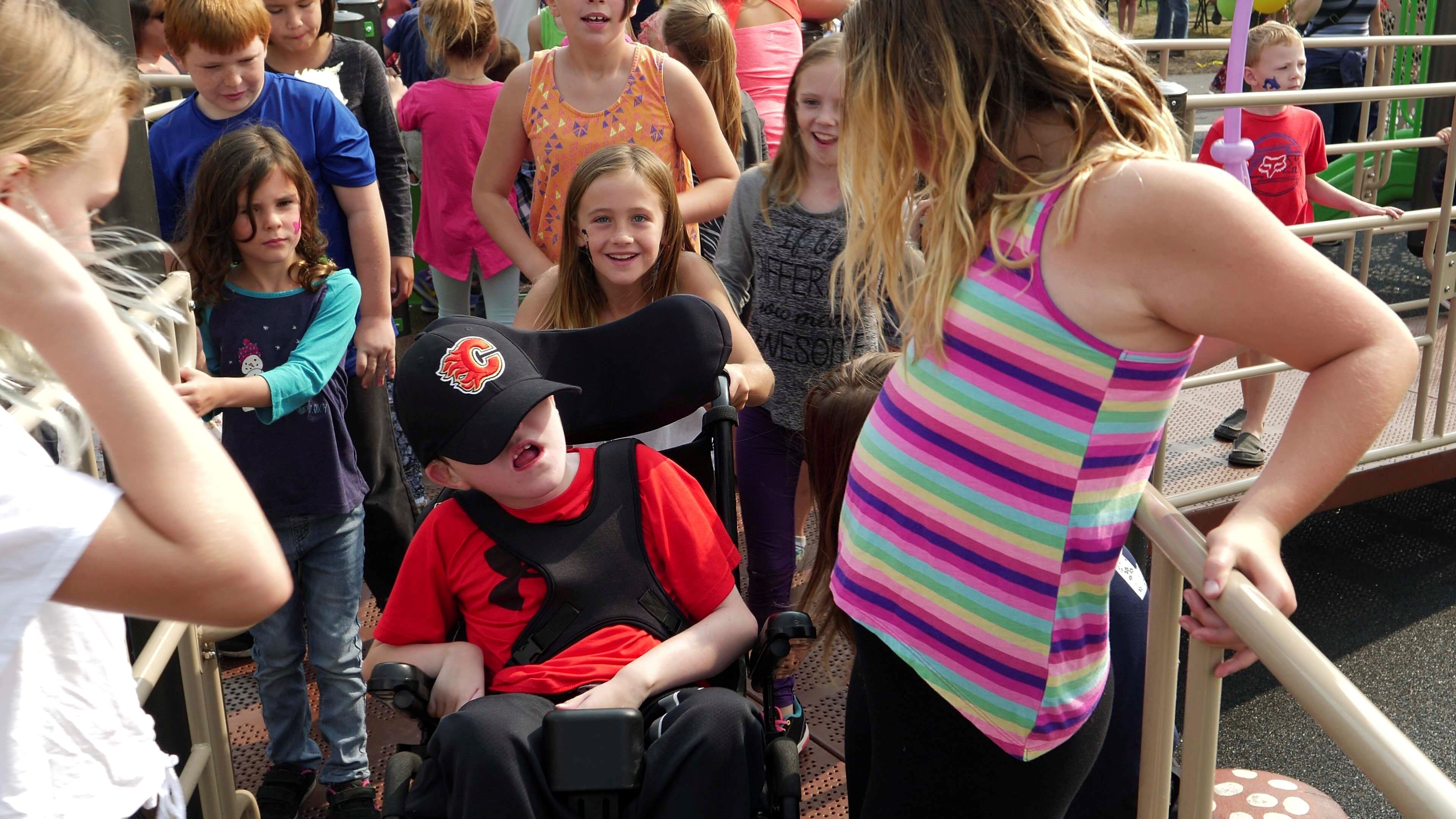Busy playground ramp crowded with children gathered around a child in a wheelchair wearing a black cap with a flame emblem; a striped tank top visible in the foreground.
