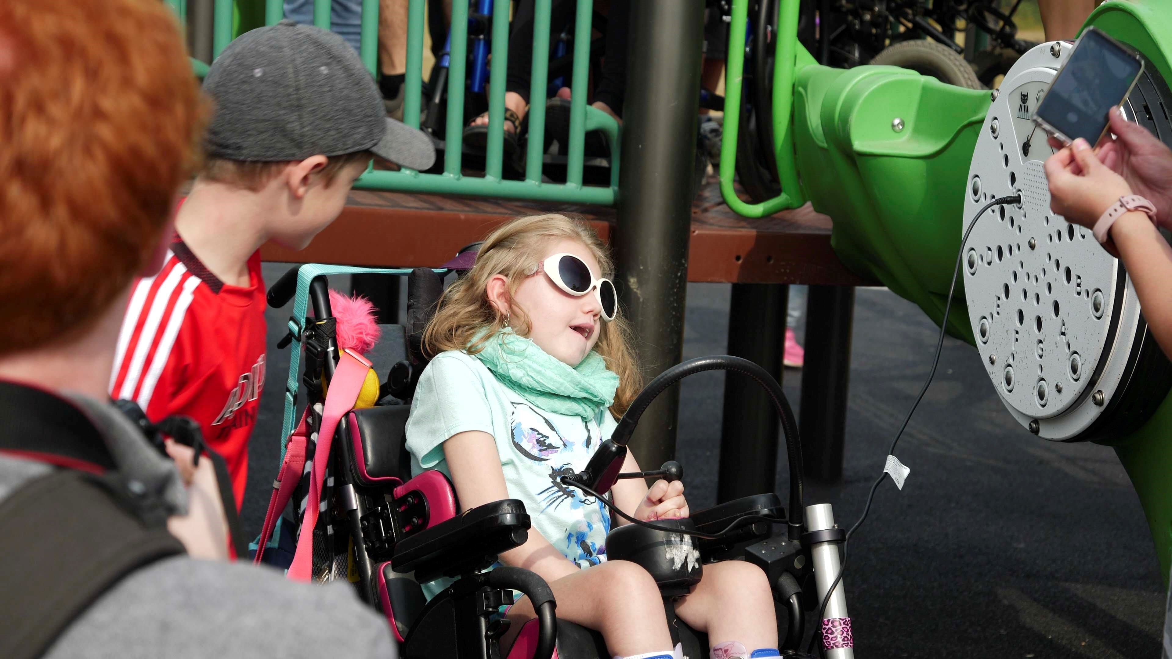 Girl wearing white sunglasses seated in a wheelchair leans toward a green play panel while a person at right holds a smartphone near the panel.
