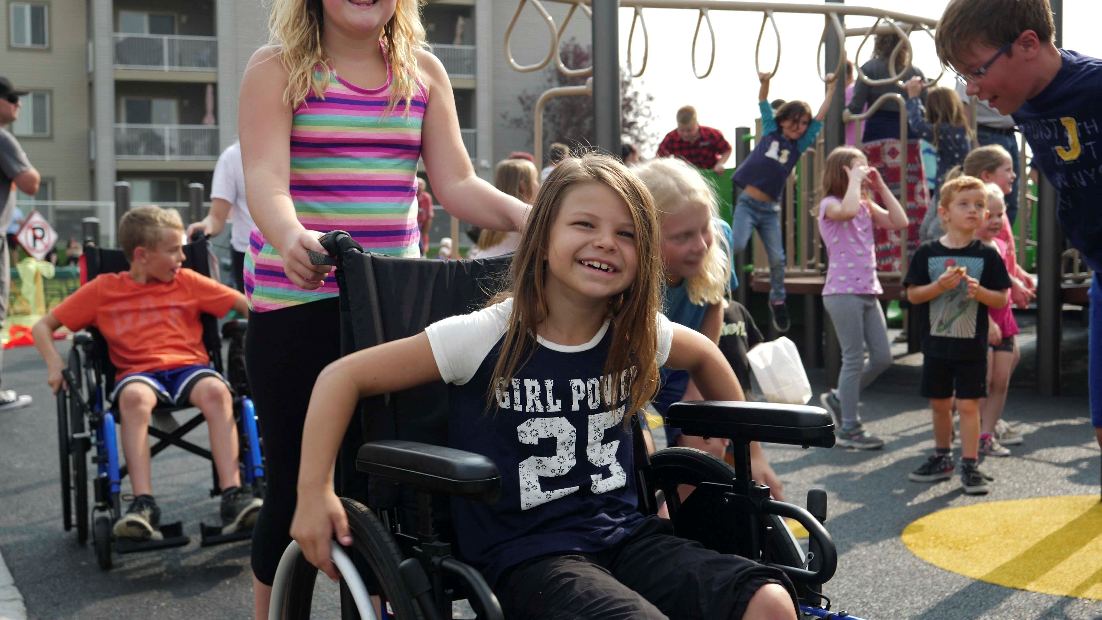 Smiling girl in a wheelchair pushed by another child, with a busy playground and many children playing in the background.