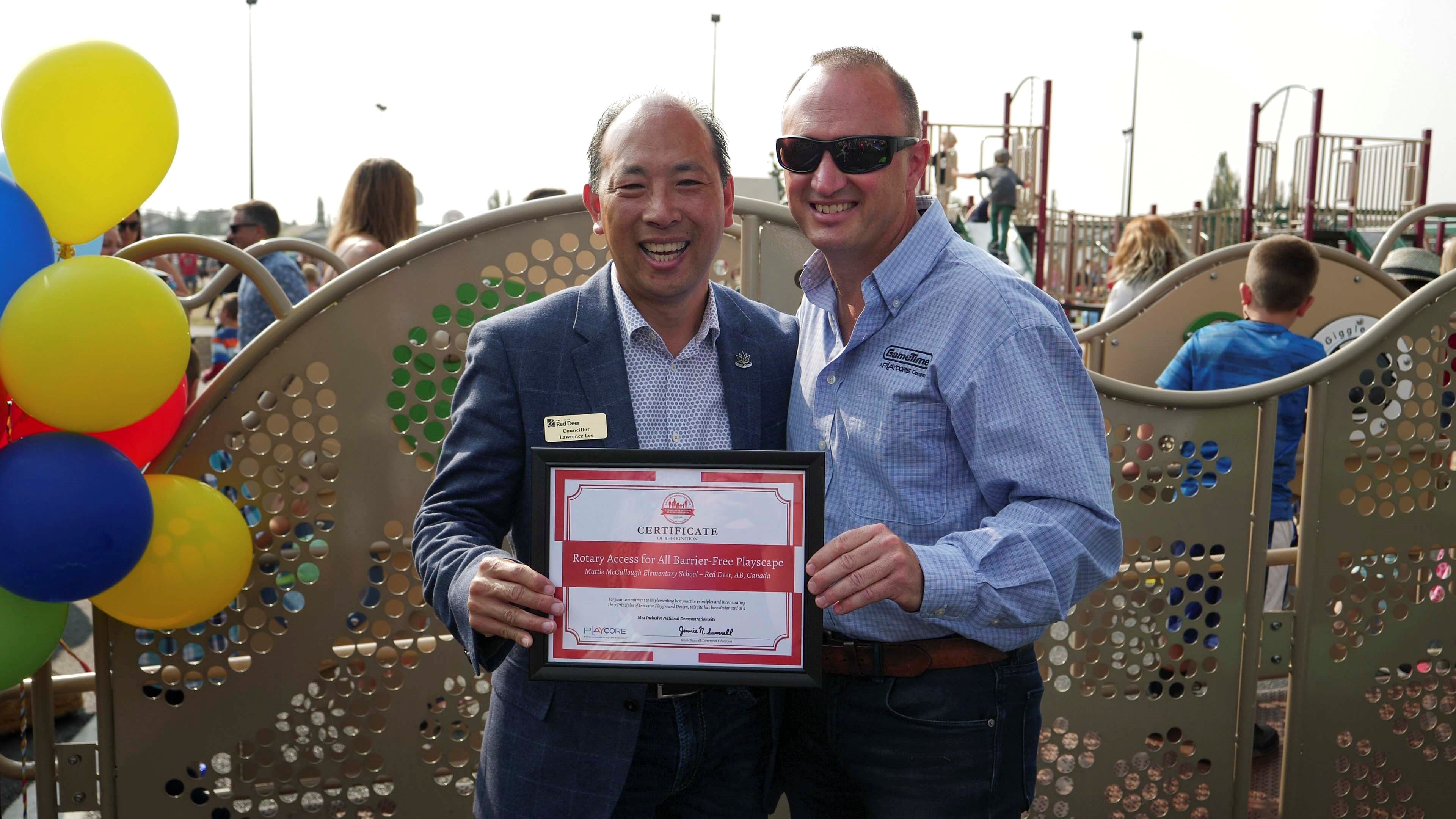 Two smiling men hold a framed certificate in front of perforated playground panels, colorful balloons at left.