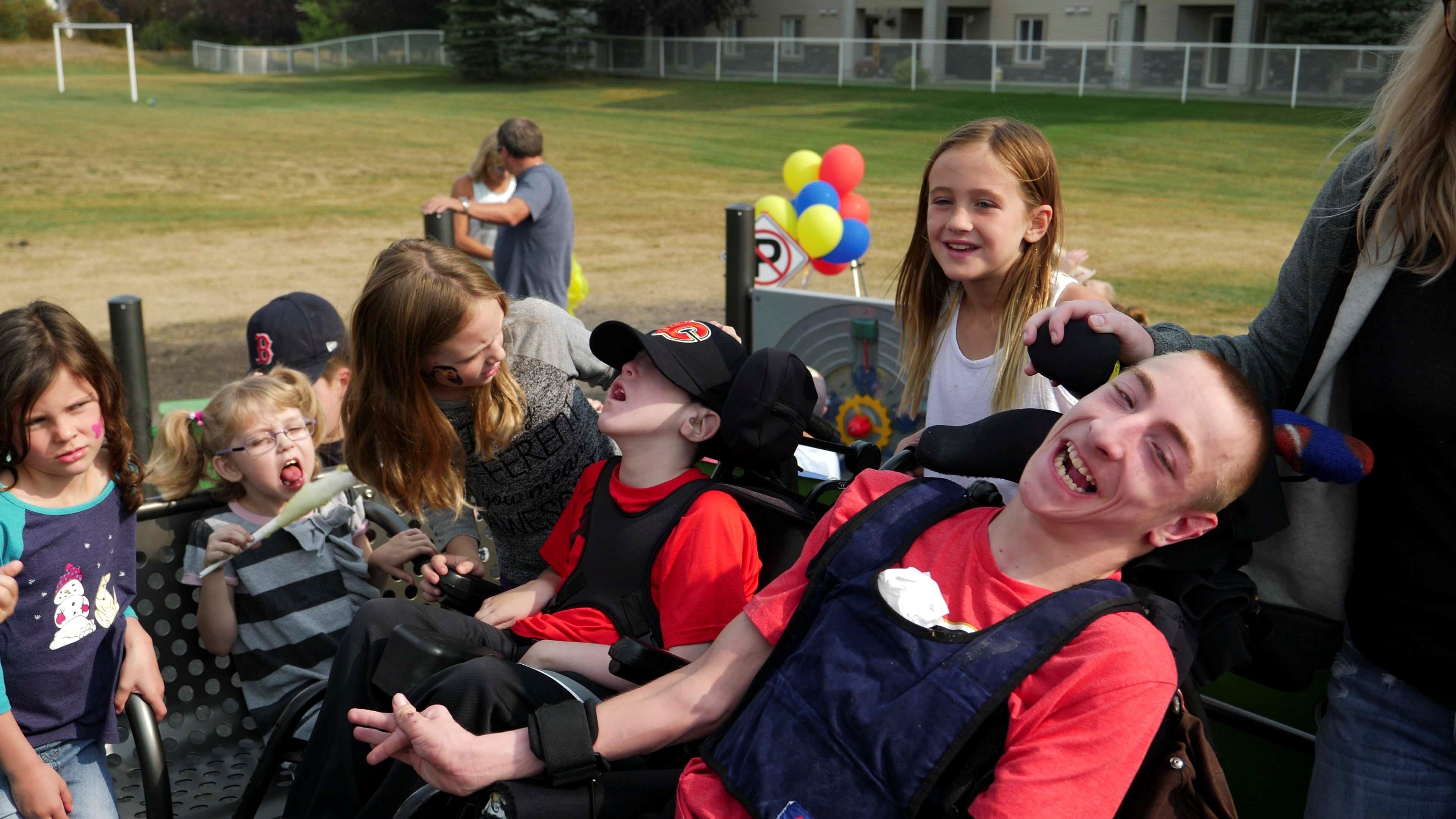 Children gathered around two boys in wheelchairs; one boy in red laughs broadly while others lean in and watch.