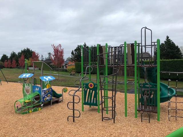 Wide view of playground showing a small play vehicle and a larger green climbing structure with spiral ladders and platforms on woodchips.