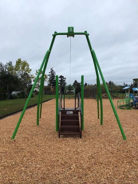 Front view of a green A-frame swing structure with central steps to a platform and an overhead cable, set on woodchips.