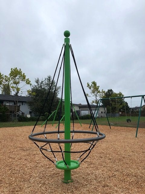 Tall green pole with suspended circular rope-basket seat held by black ropes, set on woodchip surfacing with swing frame in background.