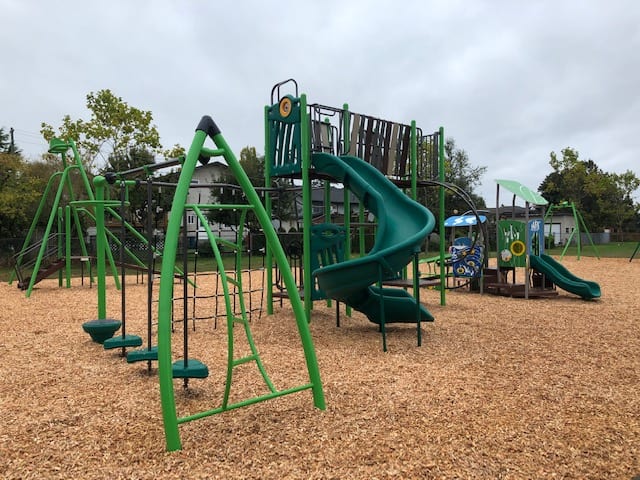 Wide view of playground showing a green arch ladder with stepping pads, central spiral slide and connected climbing platforms on woodchips.