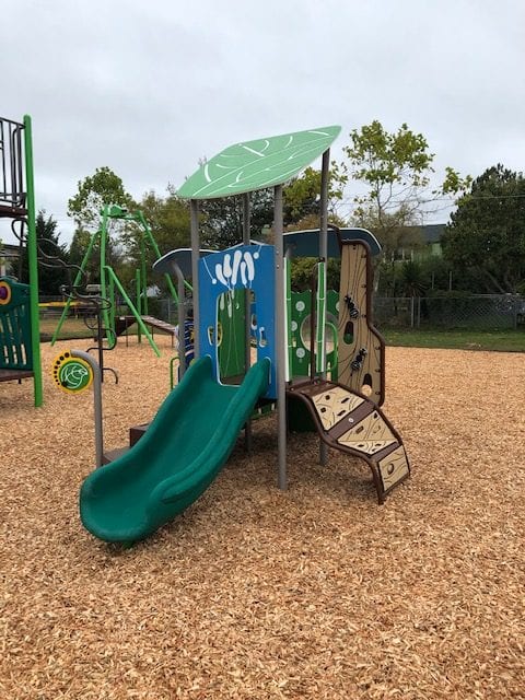 Small play structure with a green slide under a leaf-shaped canopy, decorative panels and a short curved climbing ramp on woodchips.