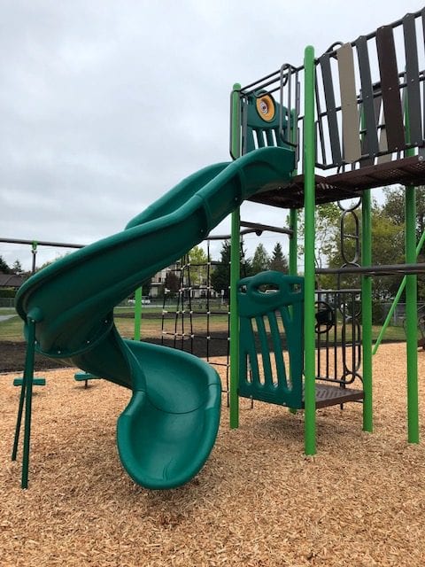 Curving green spiral slide descending from an elevated platform with green support posts over woodchip surfacing.