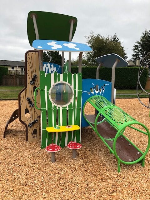 Green-and-blue play tower with round porthole, yellow bench, two red mushroom stools and green tubular crawl tunnel on woodchips.
