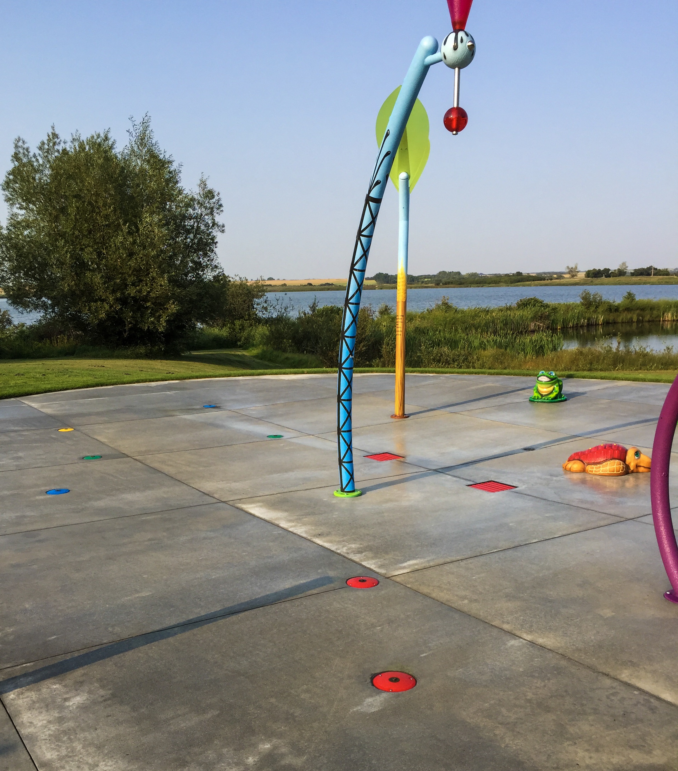 Two tall decorative poles—one blue with black pattern and one yellow with a green leaf—over a concrete pad with colored floor nozzles, a frog seat and lake beyond.
