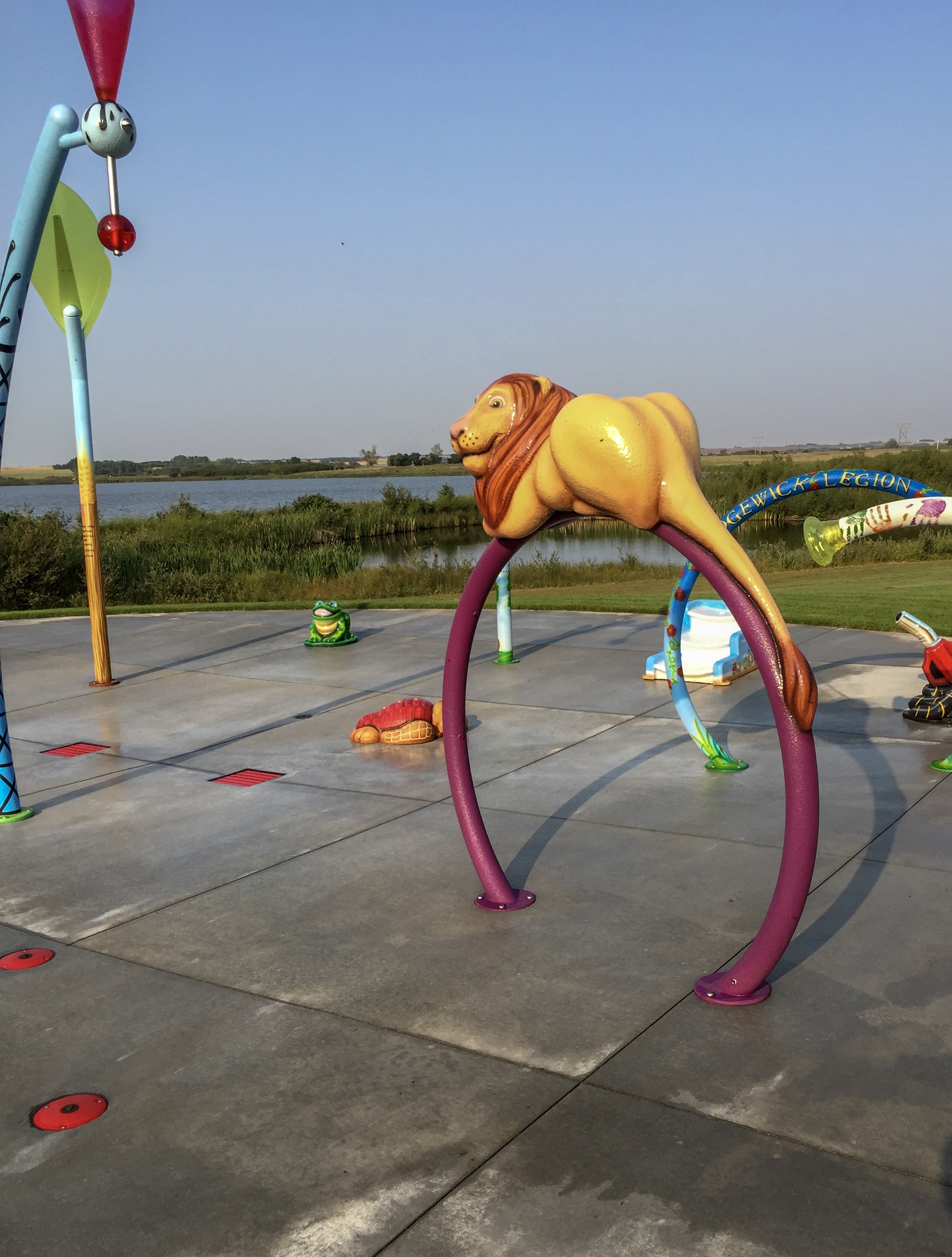 Yellow lion sculpture atop a purple circular arch on a concrete splash pad, with a green frog, turtle, colorful poles and a lake beyond.
