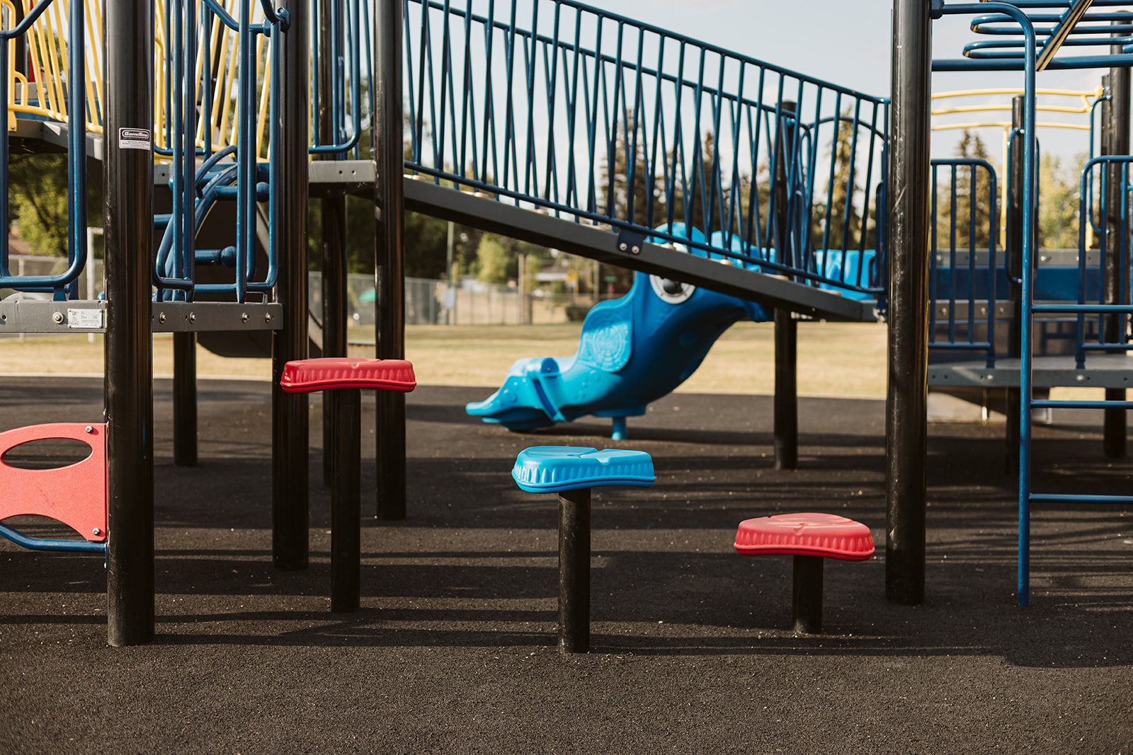 Row of low red and blue stepping pads beneath a raised play platform, with a blue molded slide and black support posts, grass and fence beyond.
