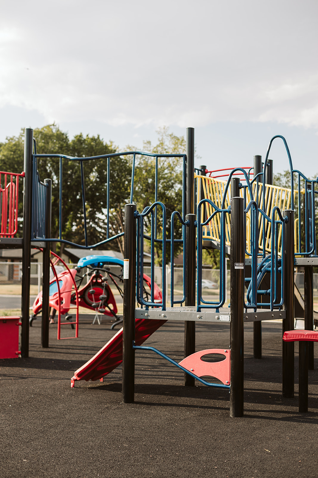 Vertical view through playground platforms showing black posts, blue and yellow railings, a red climbing pod and a small red slide on rubber surfacing.