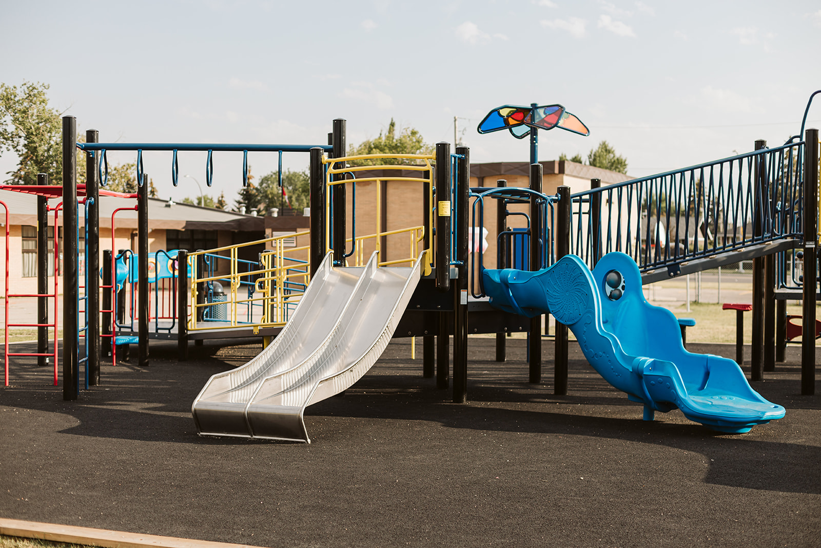 Twin straight metal slides at center with a blue curved slide to the right, black posts and yellow railings on a playground.