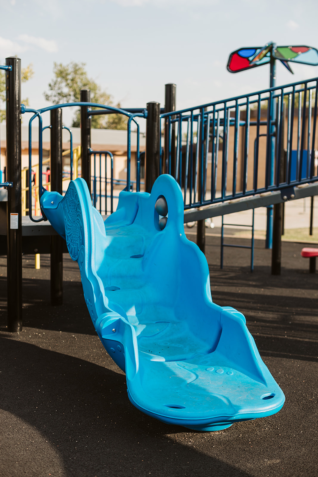 Close-up of a long blue molded multi-seat slide leading down from a raised platform, with railings and other playground structures behind.