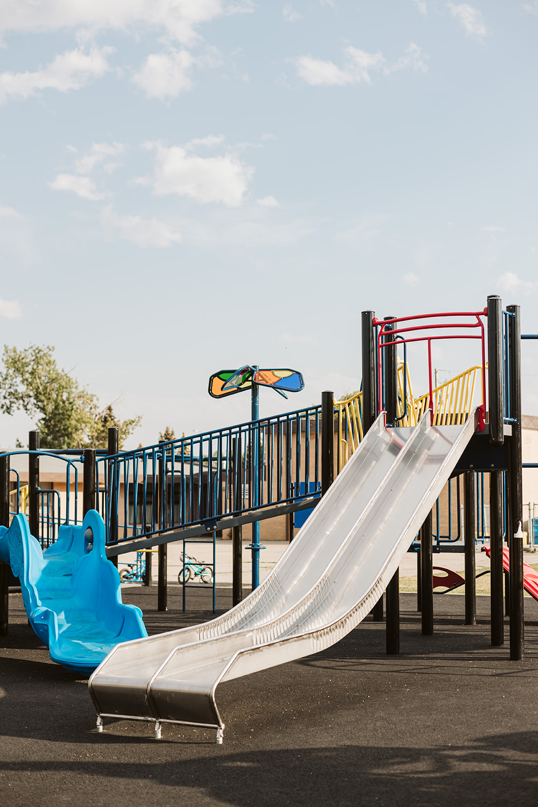 Two parallel metal slides beside a blue molded wave slide, with a colorful windmill-like spinner and a school building in the background.