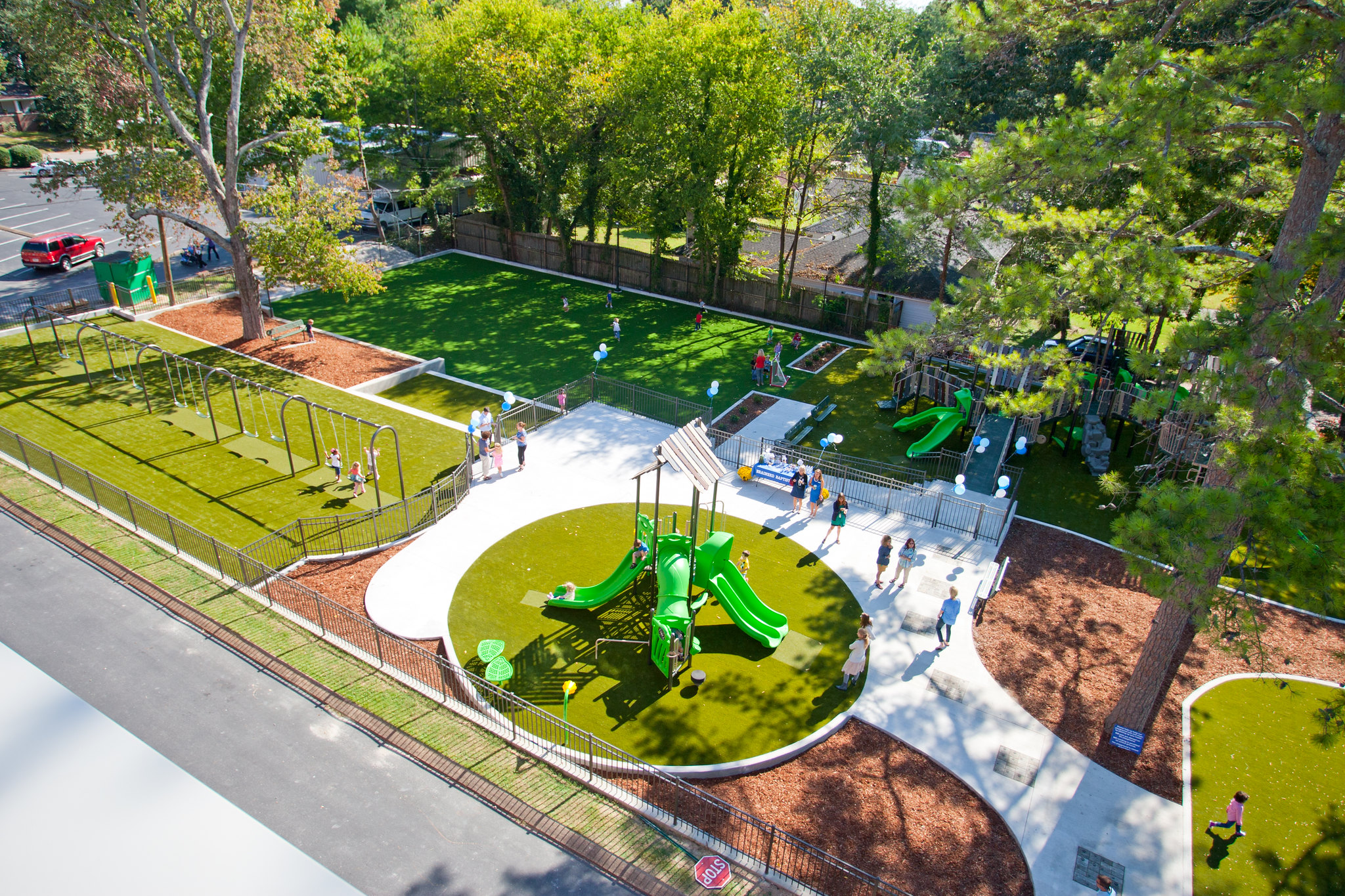Aerial view of a fenced community playground with bright green slides, swings, artificial turf and children playing under trees