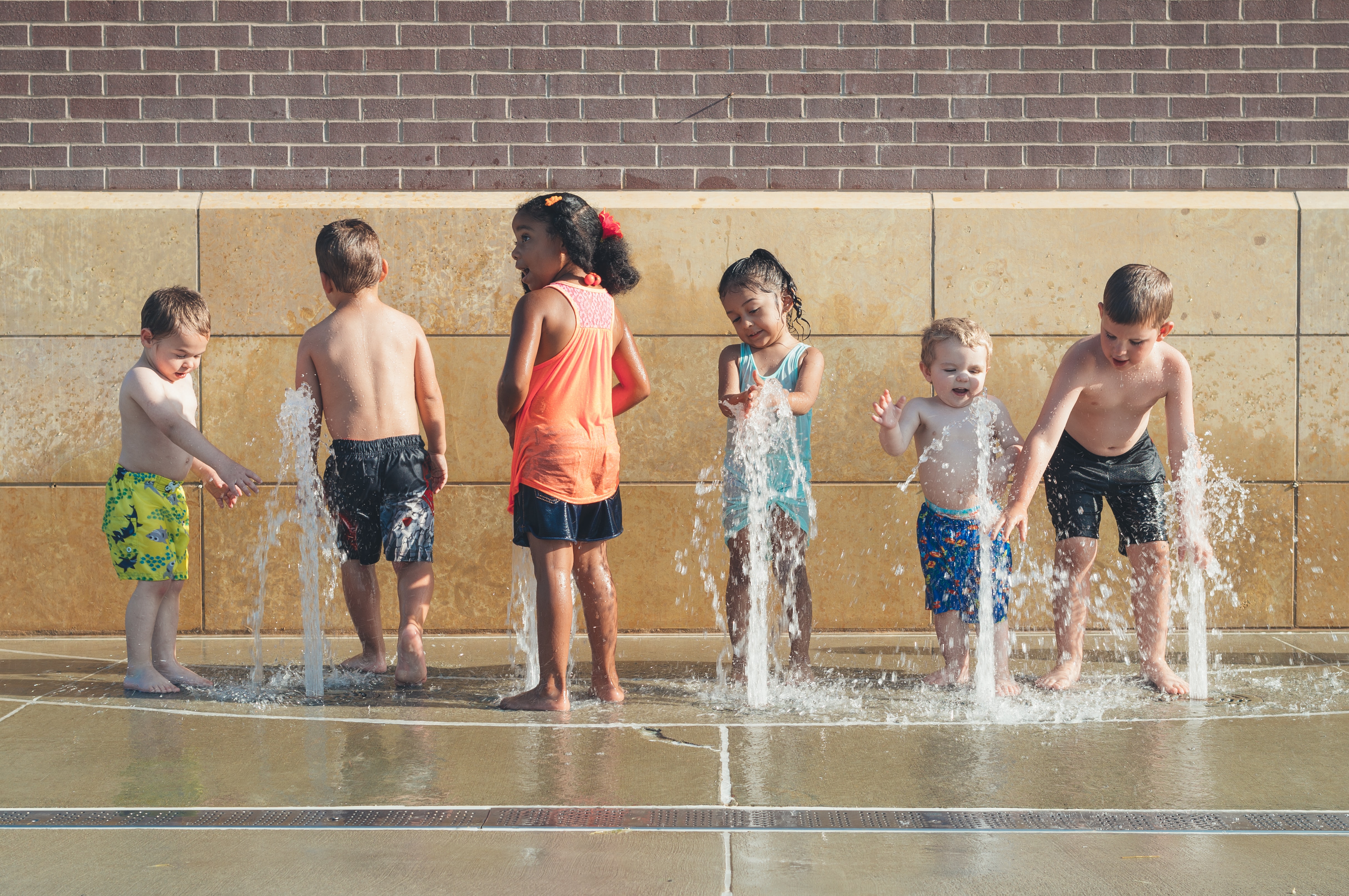 Six children playing in vertical water jets at a splash pad against a brick and stone wall