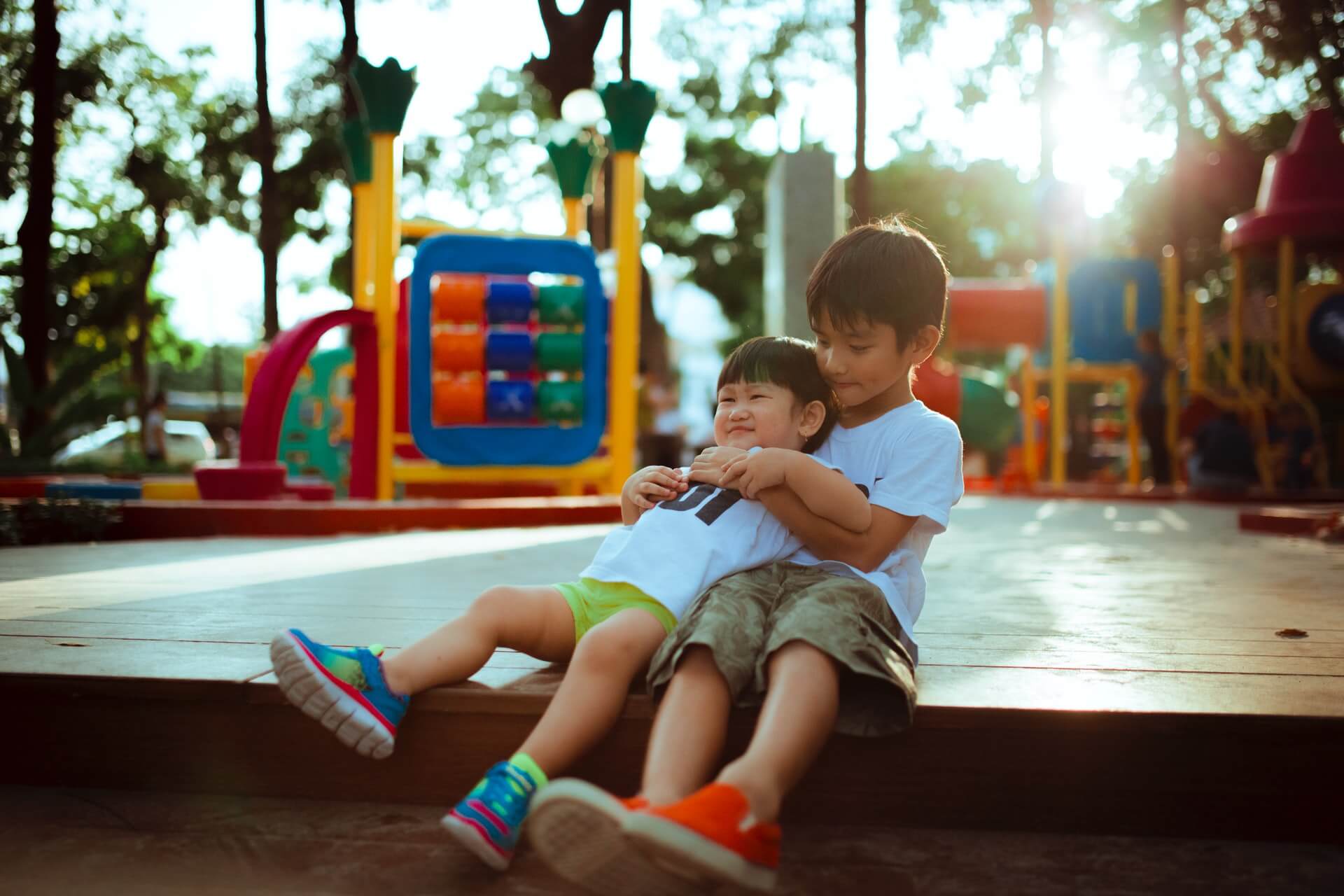 Older child hugs a younger child as they sit on a playground platform with colorful equipment and backlit sunlight