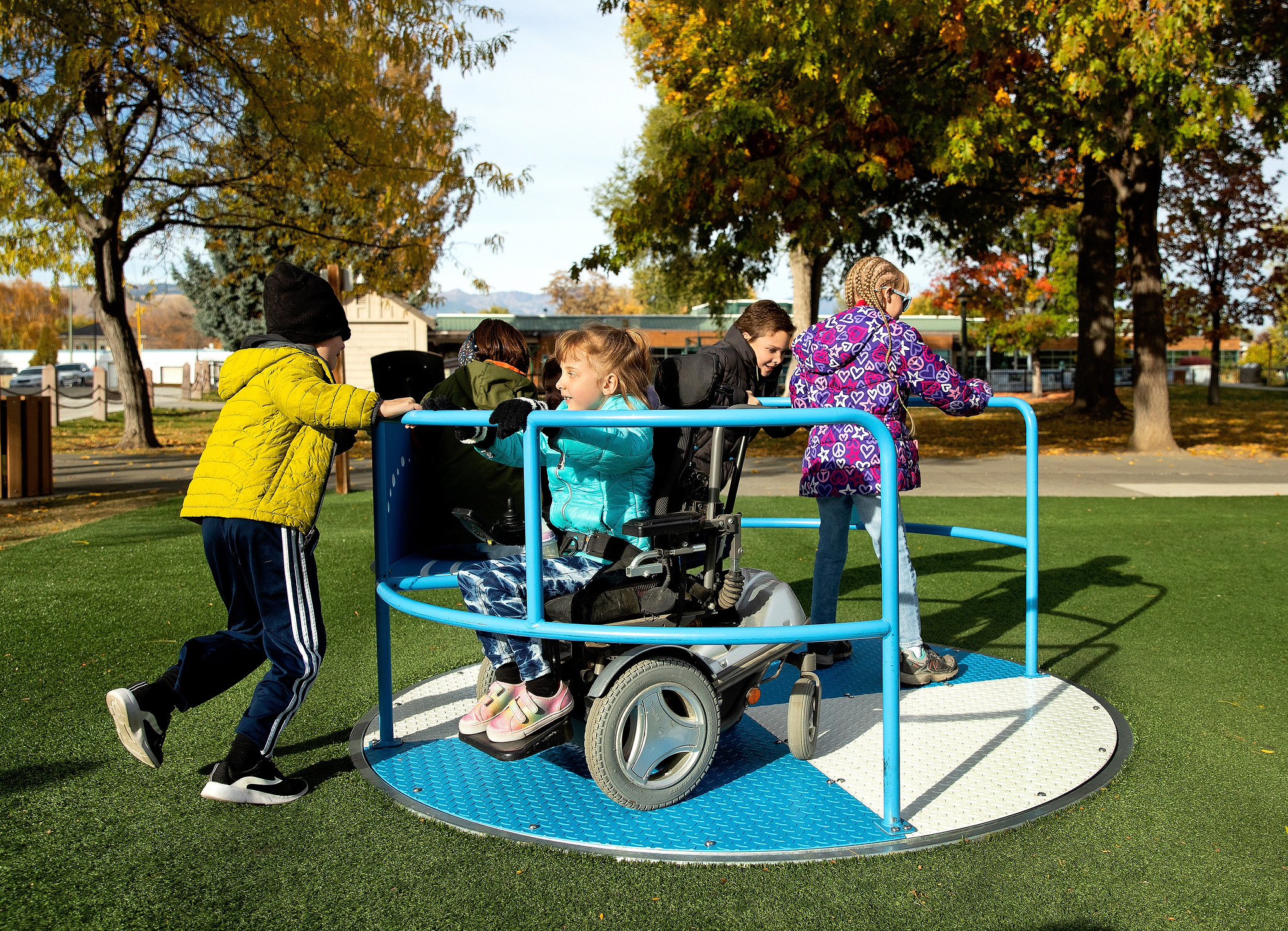 Children push and ride a blue accessible playground spinner, one child seated in a power wheelchair, with fall trees and park paths visible