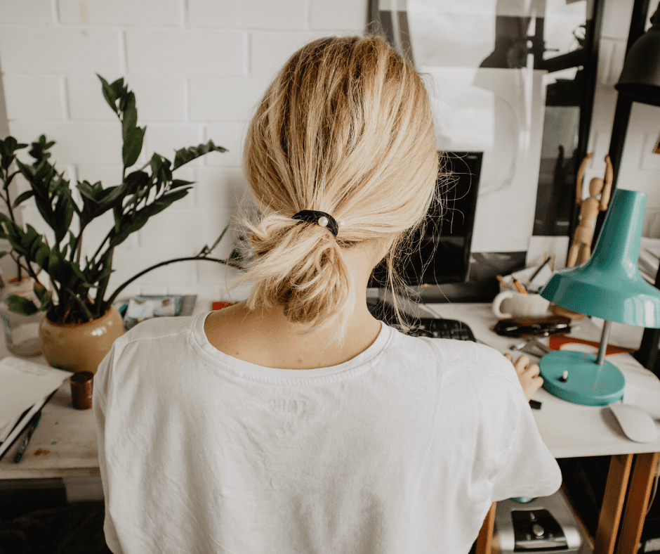 Person with blonde hair in a low bun seated at a home desk, typing on a computer with a potted plant and teal desk lamp nearby