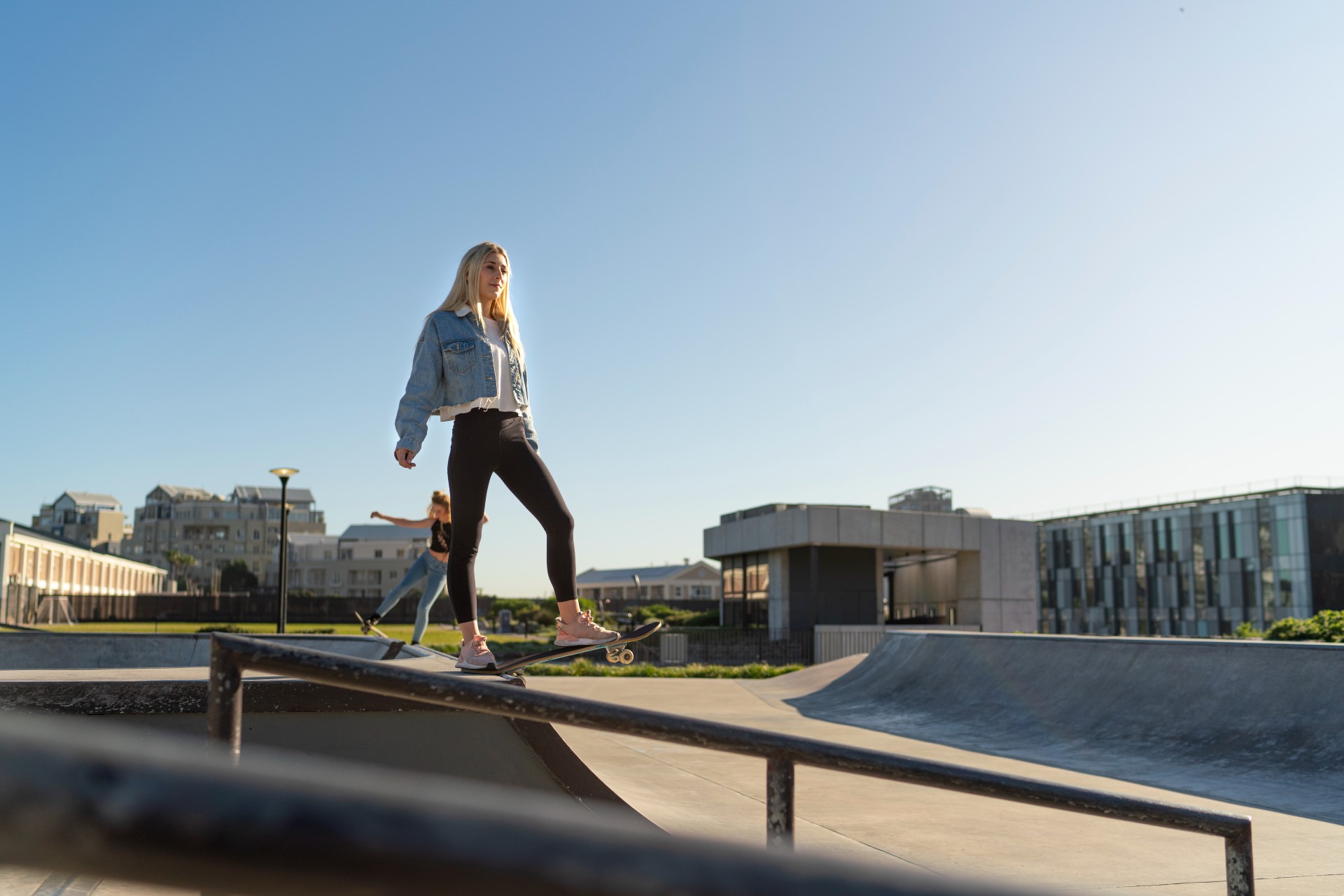 Person balancing on a skateboard along a metal rail at an outdoor concrete skatepark with buildings behind