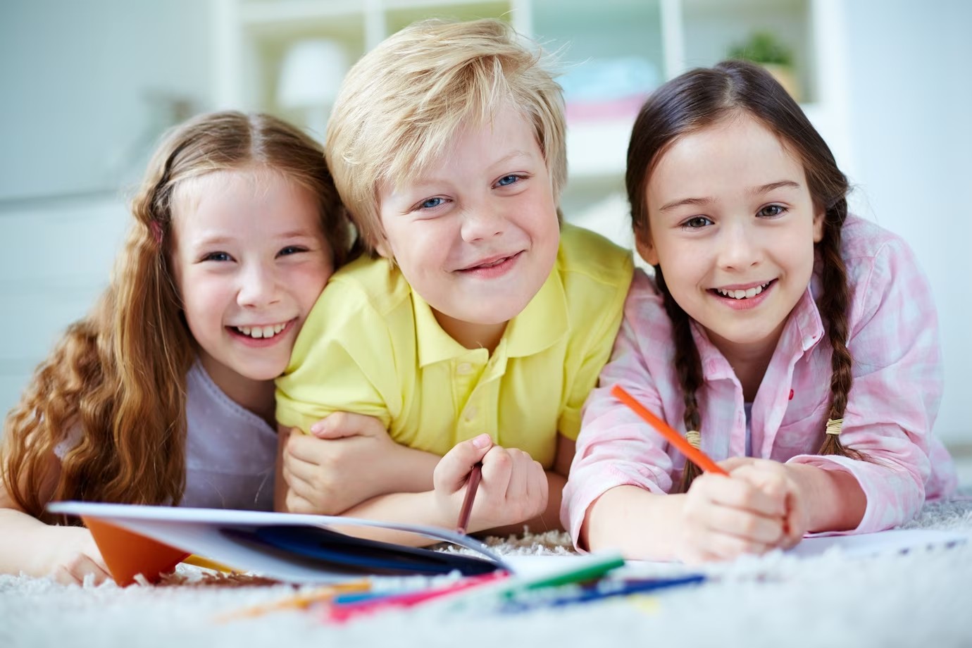 Three smiling children lying on a carpet, drawing on paper with colored pencils