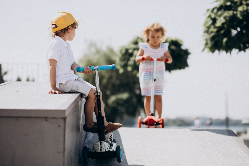Young boy in a yellow cap sitting on a skatepark ledge with a scooter as a girl in a striped dress scoots toward him