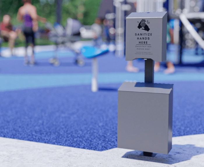 Hand sanitizer dispenser on a post at a playground; sign reads 'SANITIZE HANDS HERE', blurred children and blue play surface behind