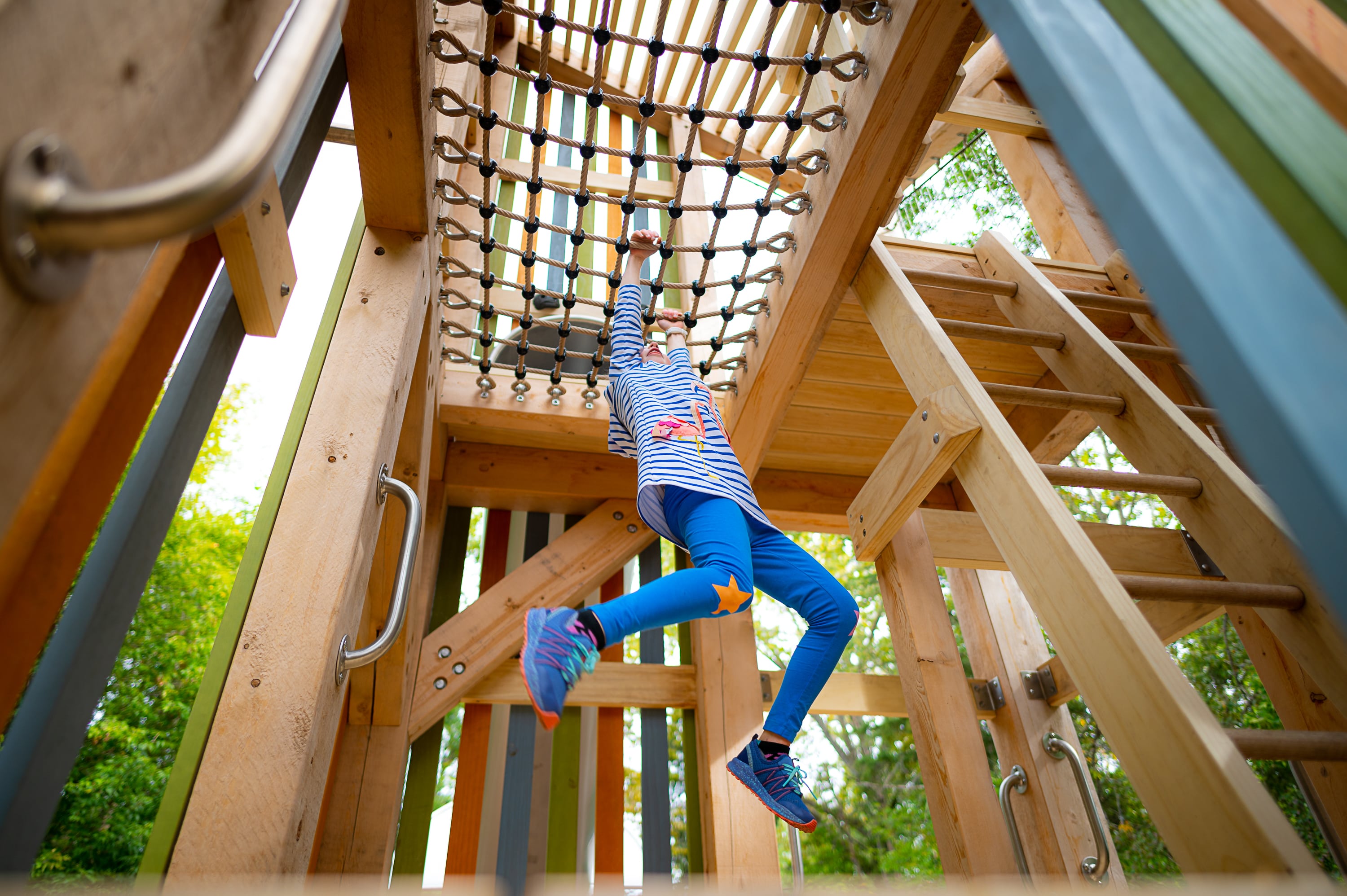 Child in striped shirt and blue leggings hanging from an overhead rope net inside a wooden playground structure