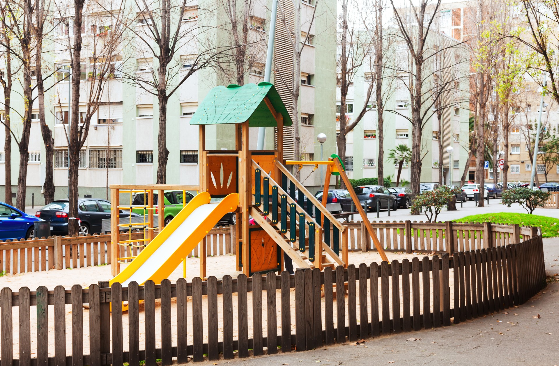 Fenced neighborhood playground with wooden picket fence surrounding a wooden play structure with yellow slide and green roof