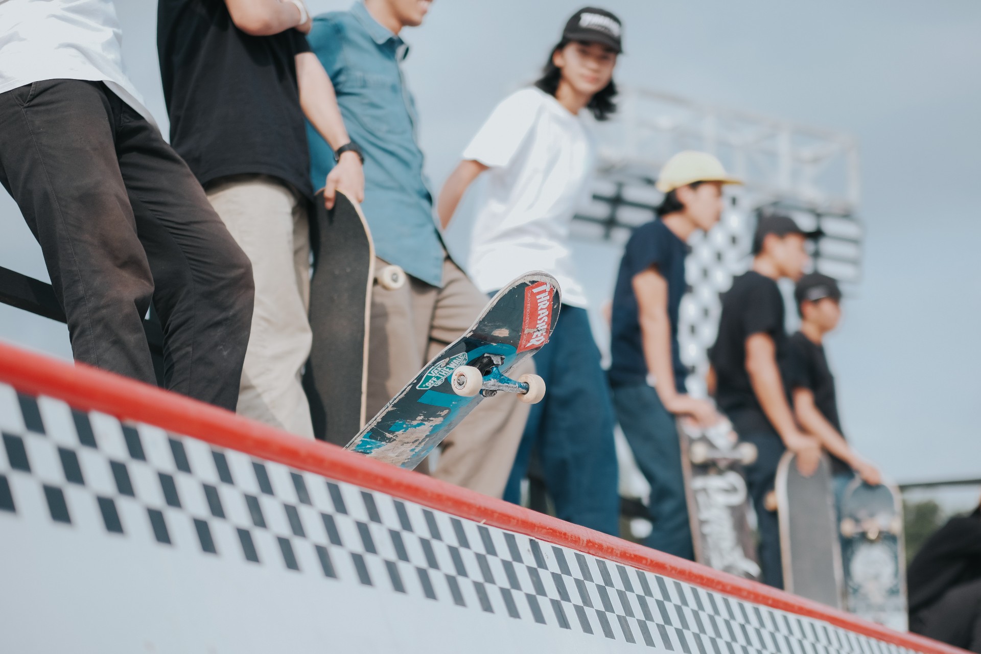 Skateboarders lined up along a checkerboard-pattern ramp edge, a worn skateboard with a red "THRASHER" sticker in the foreground