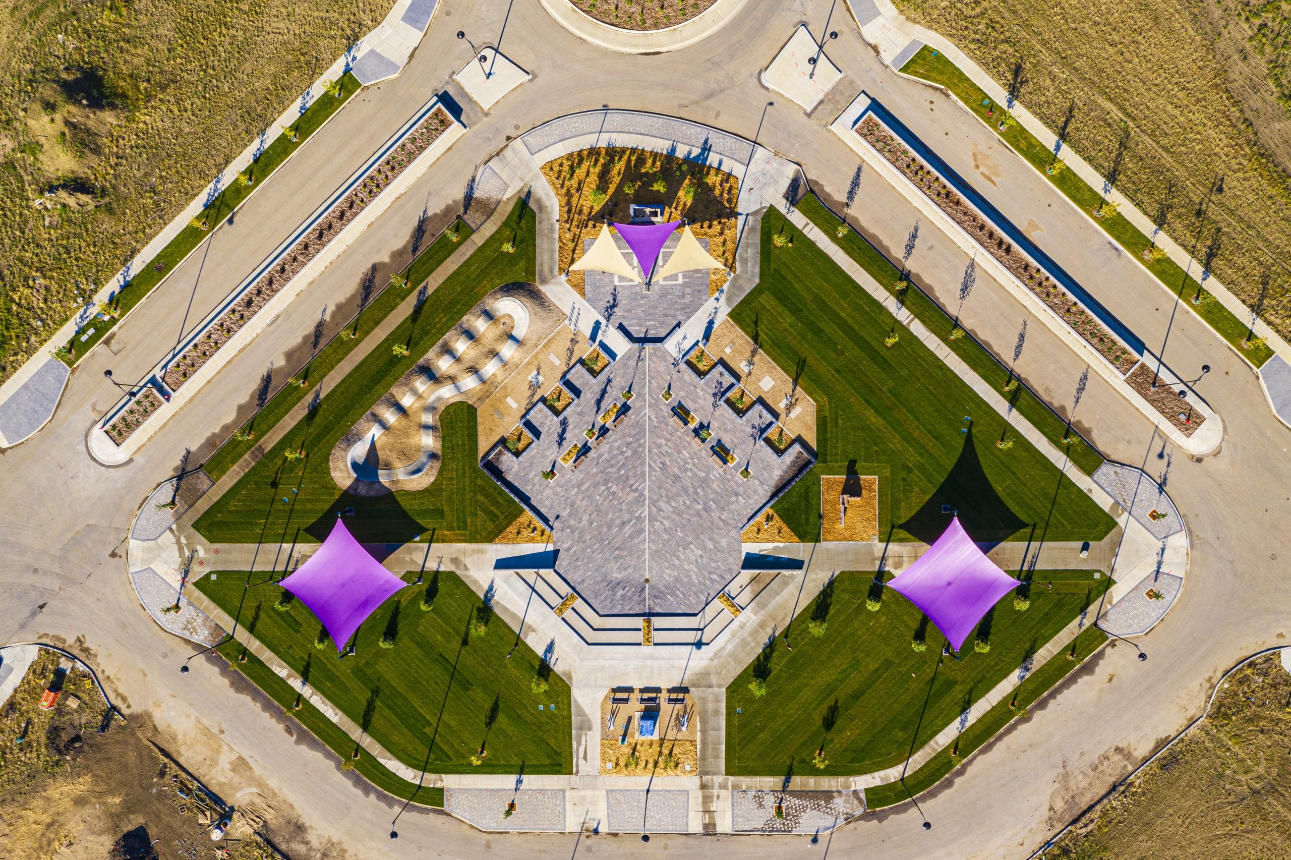 Aerial view of a symmetrical neighborhood park with central paved plaza, pathways, green lawns and three purple shade sails