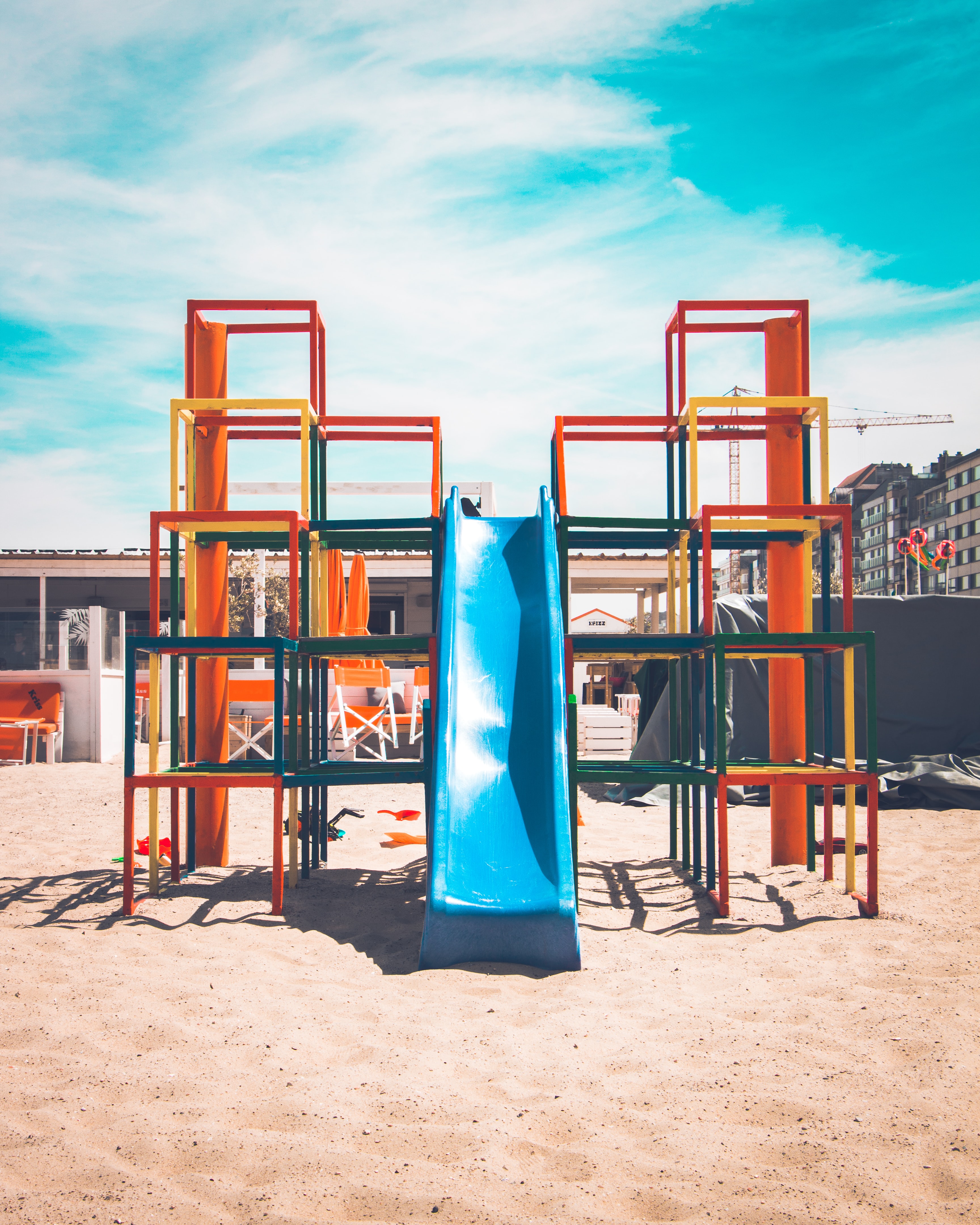 Colorful stacked square-frame playground structure with a central blue slide on sand under a bright sky