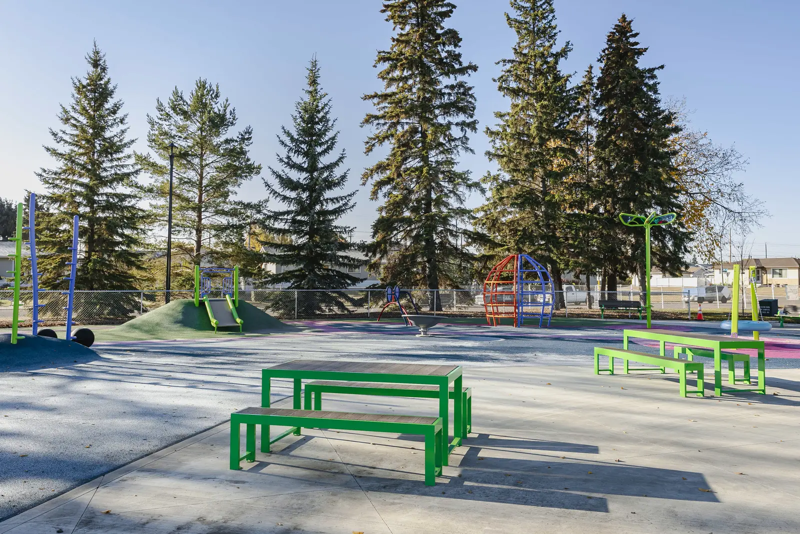 Green picnic tables on the plaza, a slide mound, dome climber and leaf-topped post beyond