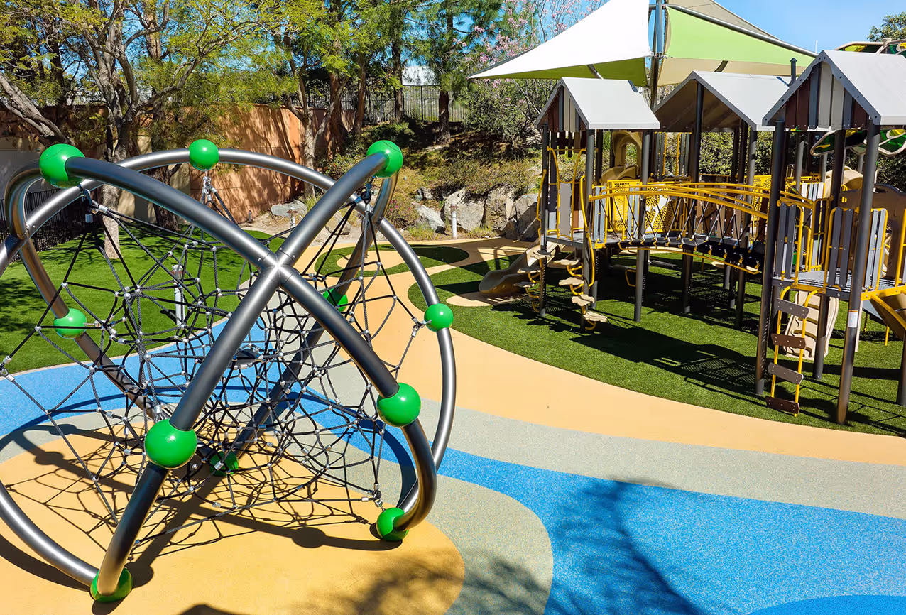 Modern playground with climbing sphere, vibrant blue and beige flooring, shaded play area with slides and bridges, surrounded by greenery.