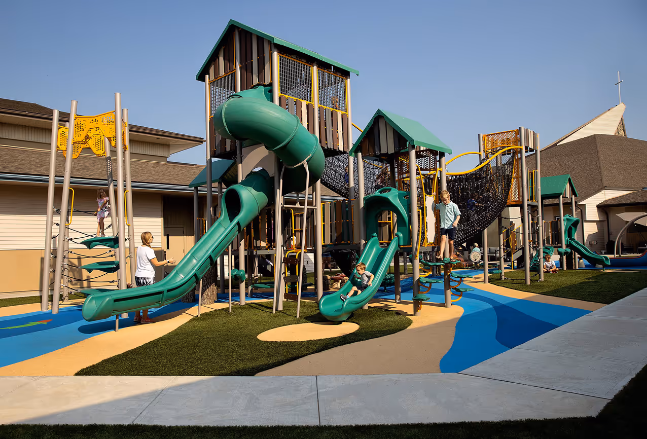 Playground with a climbing sphere and a structure with slides and bridges on colorful soft flooring, surrounded by trees under a sunny sky.